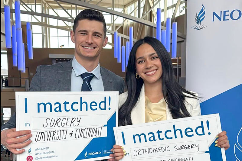 match-day-2026-800x533 Two fourth-year medical students hold signs showing where they will be going for residency.