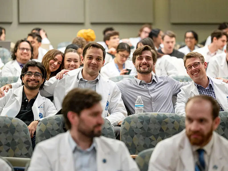 Northeast Ohio Medical University A group of medical students, many wearing white coats, sit together in a lecture hall smiling and talking before class, creating a collegial atmosphere.
